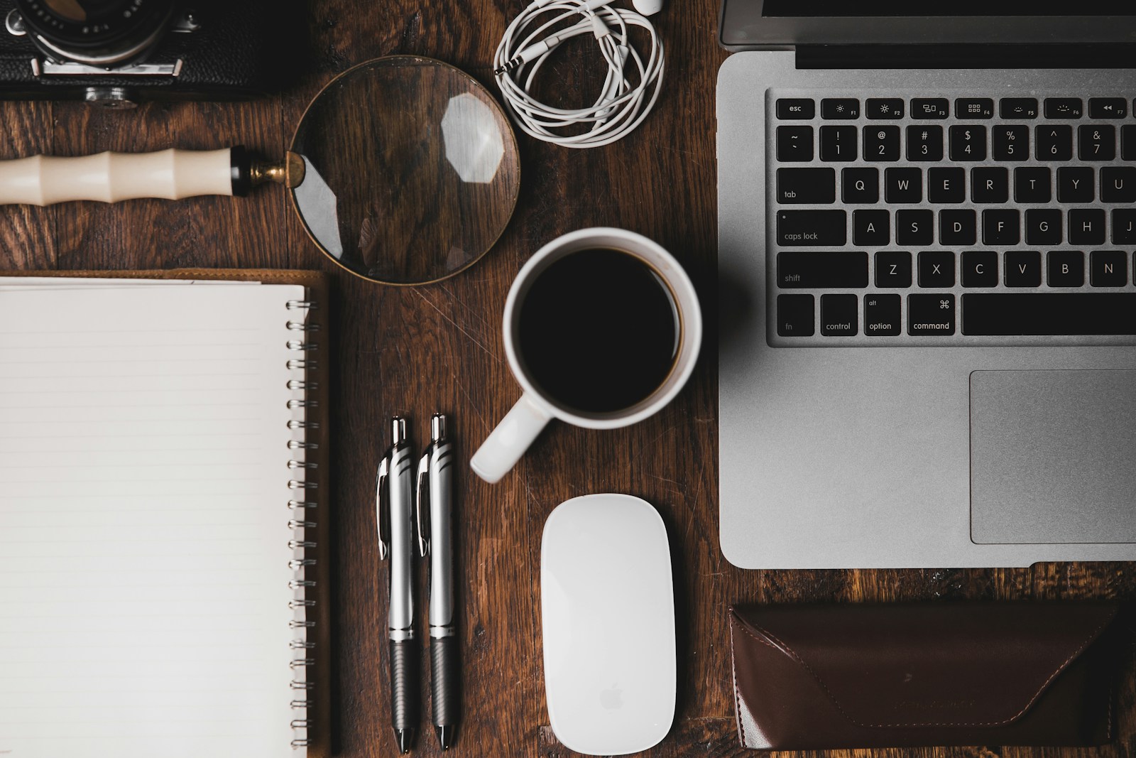 Varmt arbeidsbord i mørkt tre med laptop, kaffekopp, notatbok og kamera sett ovenfra